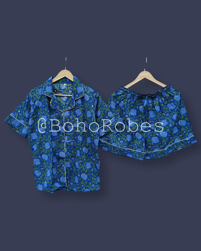 Puede incluir: Conjunto de pijama con estampado floral azul. El conjunto incluye una camisa de manga corta con botones y pantalones cortos a juego, ambos con ribete blanco. La tela presenta un patr&oacute;n repetido de flores azules y hojas verdes. El texto "@BohoRobes" es visible.