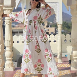 May include: A white maxi dress with a pink and green floral print. The dress has a gathered waist and long sleeves. The model is wearing the dress with sandals and a pair of silver anklets.
