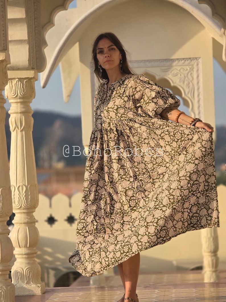 May include: A long, flowing dress with a floral pattern in olive green and cream. The dress has a loose, comfortable fit with short sleeves and a gathered waist. The text "@BohoRobes" is visible on the dress.