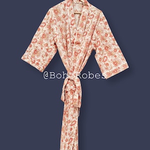 Puede incluir: Bata con estampado floral crema y coral colgada de una percha de madera. La bata presenta un patrón repetido de flores y hojas. Tiene mangas anchas y un cinturón a juego. El texto "@Bohorobes" está en la parte inferior.