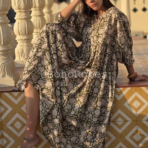 May include: A long, flowing dress with an olive green and cream floral pattern. The dress has long sleeves and a relaxed fit. The text "@BohoRobes" is visible on the garment. The dress is worn outdoors.