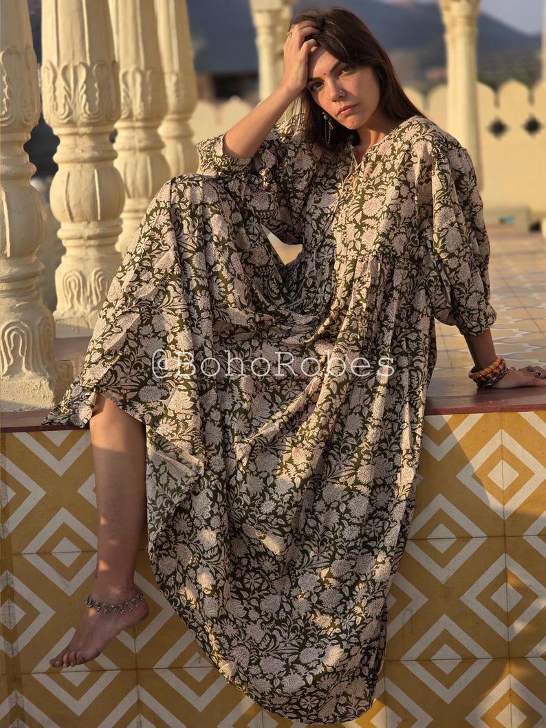 May include: A long, flowing dress with an olive green and cream floral pattern. The dress has long sleeves and a relaxed fit. The text "@BohoRobes" is visible on the garment. The dress is worn outdoors.