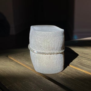 May include: A white velvet ring box with a silver beaded bracelet on top.