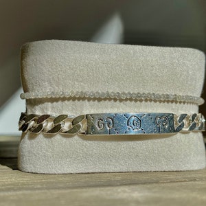 May include: A silver chain bracelet with a silver rectangular tag engraved with the letters "GG" and a skull design. The bracelet is displayed on a white velvet cushion.