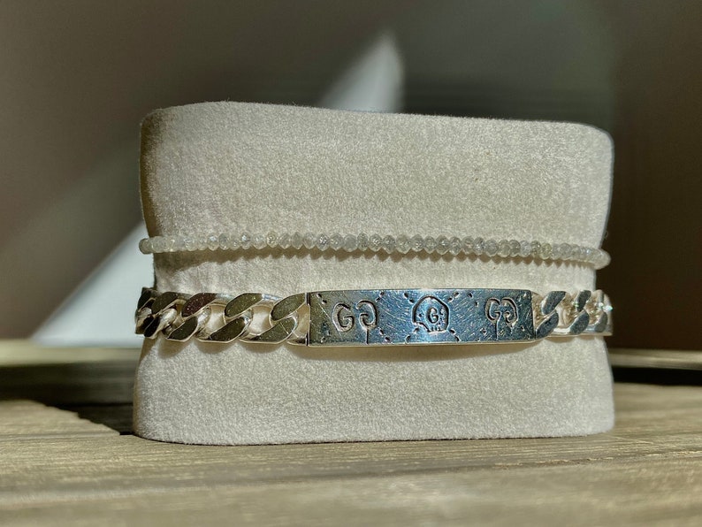 May include: A silver chain bracelet with a silver rectangular tag engraved with the letters "GG" and a skull design. The bracelet is displayed on a white velvet cushion.