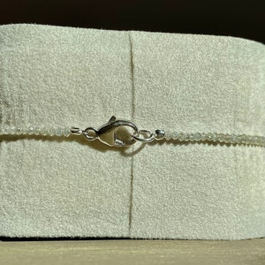 May include: A silver clasp closure on a white beaded necklace. The necklace is displayed on a white velvet cushion.