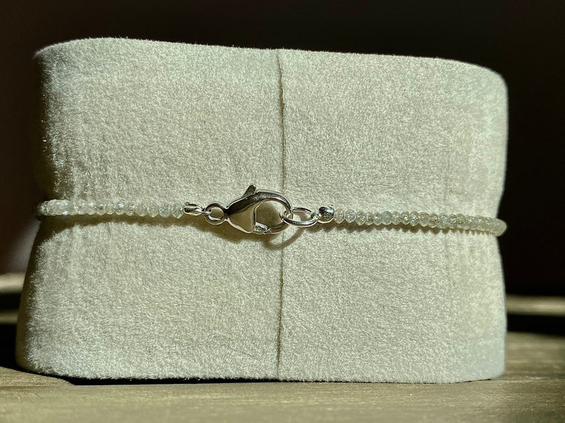 May include: A silver clasp closure on a white beaded necklace. The necklace is displayed on a white velvet cushion.