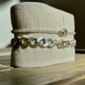 May include: Two silver chains on a white velvet display stand. The top chain is a delicate, beaded chain. The bottom chain is a thick, curb chain.