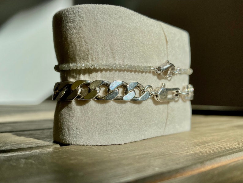 May include: Two silver chains on a white velvet display stand. The top chain is a delicate, beaded chain. The bottom chain is a thick, curb chain.