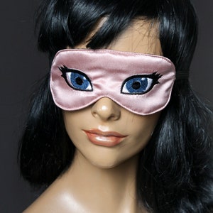 May include: A pink satin sleep mask with embroidered blue eyes and eyelashes.