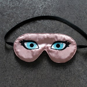 May include: A pink satin sleep mask with embroidered blue eyes and eyelashes.