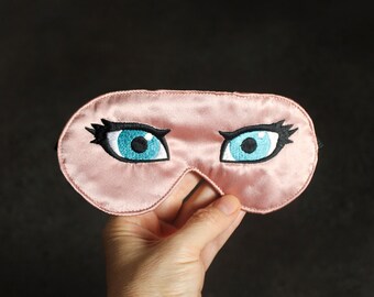 Op de afbeelding: Een roze satijnen slaapmasker met geborduurde wimpers en blauwe ogen.
