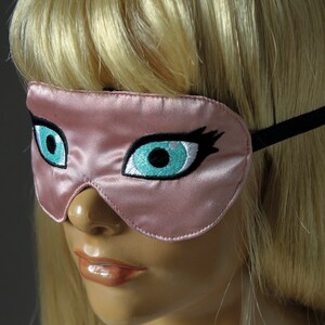 May include: A pink satin sleep mask with embroidered eyelashes and turquoise blue eyes. The mask has a black elastic band.