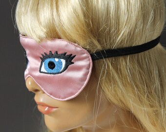Op de afbeelding: Een roze satijnen slaapmasker met een zwart elastiek. Het masker heeft een geborduurd blauw oog met zwarte wimpers.