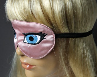 Op de afbeelding: Roze satijnen slaapmasker met een groot geborduurd oogontwerp. Het oog is blauw met zwarte wimpers en een zwarte omtrek.