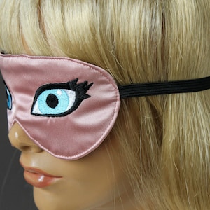 May include: A pink satin sleep mask with a black elastic band. The mask features an embroidered eye with blue and black details.