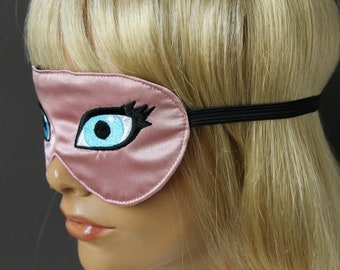 Op de afbeelding: Een roze satijnen slaapmasker met een zwart elastiek. Het masker heeft een geborduurd oog met blauwe en zwarte details.