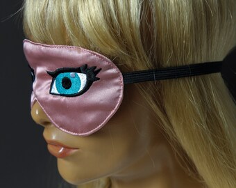 Op de afbeelding: Een roze satijnen slaapmasker met een geborduurd oogontwerp. Het oog heeft een helderblauwe iris, witte highlights en zwarte wimpers en wenkbrauwen. Het masker heeft een zwarte elastische band.