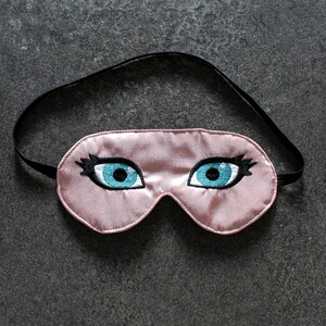 May include: A pink satin sleep mask with embroidered eyelashes and blue eyes.