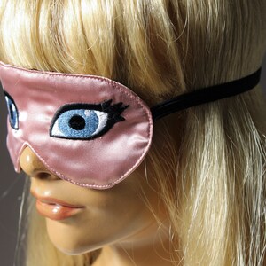 May include: A pink satin sleep mask with embroidered blue eyes and eyelashes. The mask has a black elastic band.