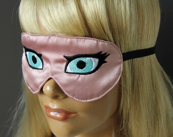 Op de afbeelding: Een roze satijnen slaapmasker met geborduurde wimpers en turquoise blauwe ogen. Het masker heeft een zwart elastiek.