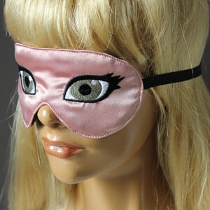 May include: A pink satin sleep mask with embroidered eyes and eyelashes. The mask has a black elastic band.