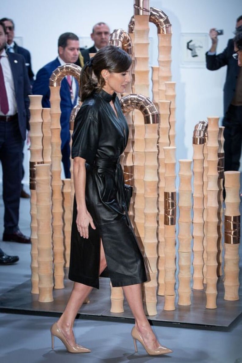 May include: A woman wearing a black leather dress with a belt and a slit up the side. She is wearing nude heels and walking past a sculpture made of stacked brown cylinders.