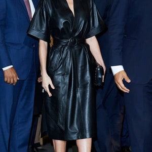 May include: A woman wearing a black leather dress with a belt and a black clutch. She is wearing nude heels and has her hair pulled back.