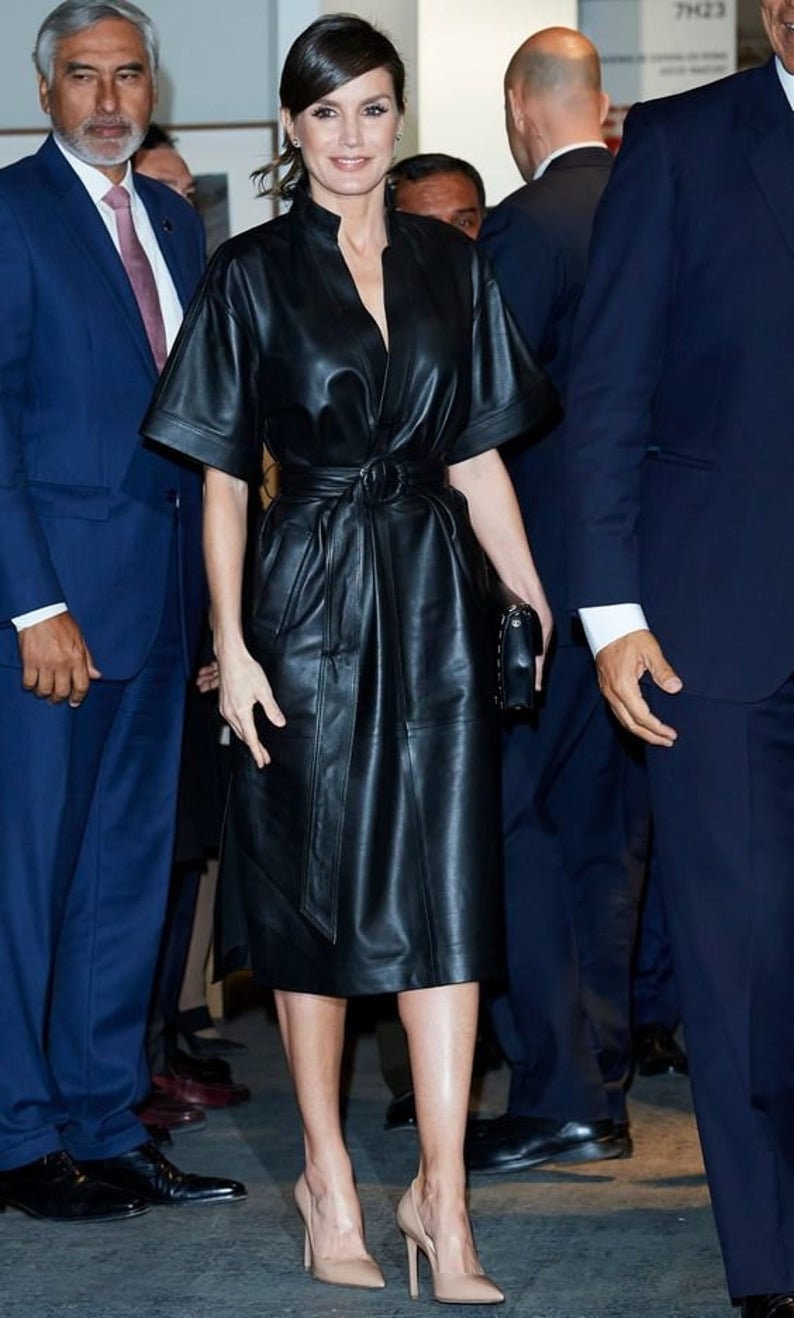May include: A woman wearing a black leather dress with a belt and a black clutch. She is wearing nude heels and has her hair pulled back.