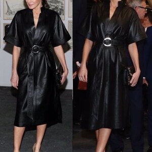 May include: A woman wearing a black leather dress with a silver belt and matching black heels. She is standing in front of a wall with artwork.
