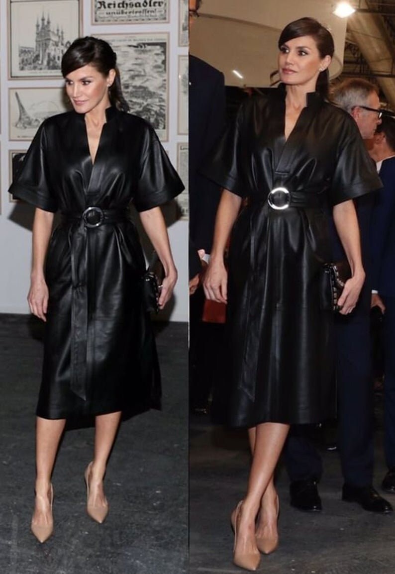May include: A woman wearing a black leather dress with a silver belt and matching black heels. She is standing in front of a wall with artwork.