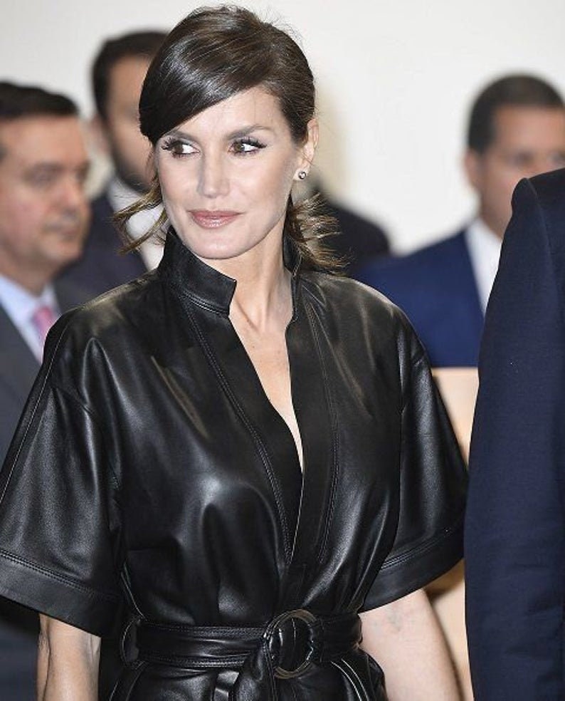 May include: A woman wearing a black leather dress with a belt. She has dark brown hair styled in a ponytail with a side part.