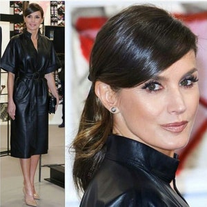 May include: A woman wearing a black leather dress with a belt. She has her hair pulled back in a ponytail with a side part.