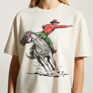 以下が含まれることがあります： クリーム色のTシャツ。グレーの馬に乗ったカウボーイのグラフィックが描かれています。カウボーイは赤いシャツ、緑のズボン、赤い帽子を着用しています。馬はギャロップしており、デザインはヴィンテージのコミックブック風です。