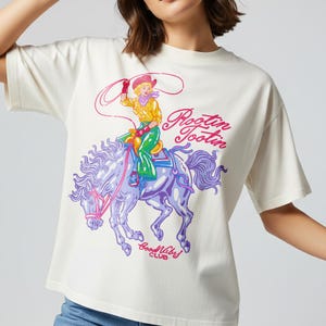 K&ouml;nnte beinhalten: Cremefarbenes T-Shirt mit einem farbenfrohen Aufdruck einer Cowgirl auf einem lila Pferd. Der Text "Rootin' Tootin' Good Vibes Club" ist in Pink und Lila aufgedruckt. Ein l&auml;ssiges Oberteil.
