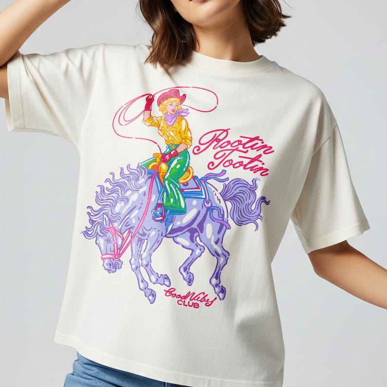 K&ouml;nnte beinhalten: Cremefarbenes T-Shirt mit einem farbenfrohen Aufdruck einer Cowgirl auf einem lila Pferd. Der Text "Rootin' Tootin' Good Vibes Club" ist in Pink und Lila aufgedruckt. Ein l&auml;ssiges Oberteil.
