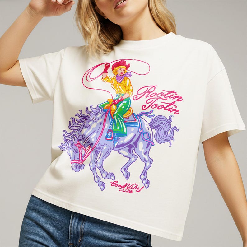 K&ouml;nnte beinhalten: Cremefarbenes T-Shirt mit einem farbenfrohen Aufdruck einer Cowgirl auf einem Pferd. Die Cowgirl tr&auml;gt ein gelbes Hemd, gr&uuml;ne Hosen und einen roten Hut. Das Pferd ist lila mit einem rosa Zaumzeug. Der Text auf dem Shirt lautet "Rootin' Tootin' Good Vibes Club."