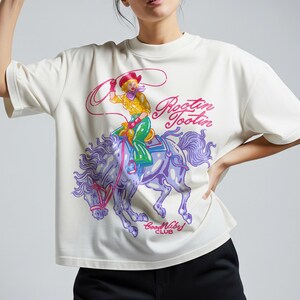 K&ouml;nnte beinhalten: Cremefarbenes T-Shirt mit einem farbenfrohen Aufdruck einer Cowgirl auf einem lila Pferd mit dem Text "Rootin' Tootin" und "Good Vibes Club". Die Cowgirl tr&auml;gt ein gelbes Hemd, gr&uuml;ne Hosen und einen roten Hut und h&auml;lt einen Lasso.