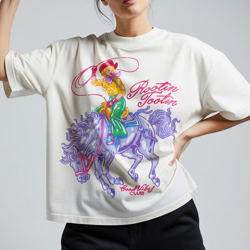 K&ouml;nnte beinhalten: Cremefarbenes T-Shirt mit einem farbenfrohen Aufdruck einer Cowgirl auf einem lila Pferd mit dem Text "Rootin' Tootin" und "Good Vibes Club". Die Cowgirl tr&auml;gt ein gelbes Hemd, gr&uuml;ne Hosen und einen roten Hut und h&auml;lt einen Lasso.