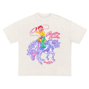 K&ouml;nnte beinhalten: Cremefarbenes T-Shirt mit einem farbenfrohen Grafikdesign einer Cowgirl auf einem Pferd mit Lasso. Der Entwurf enth&auml;lt den Text "Rootin' Tootin'" und "Good Vibes Club". Die Cowgirl tr&auml;gt einen roten Hut, ein gelbes Hemd und gr&uuml;ne Hosen.