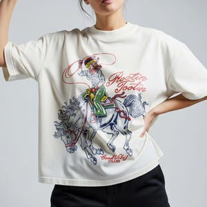 May include: Cream-colored t-shirt featuring a graphic of a cowgirl riding a horse with a lasso. The design includes the text "Rootin' Tootin'" in red script and "Good Vibes Club" in blue. The cowgirl wears a hat, green pants, and a white shirt.