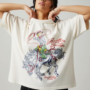 May include: Cream-colored t-shirt featuring a graphic of a cowgirl riding a horse with a lasso. The design includes the words "Rootin' Tootin" in red script and "Good Vibes Club" in blue. The shirt has short sleeves.