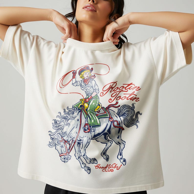 May include: Cream-colored t-shirt featuring a graphic of a cowgirl riding a horse with a lasso. The design includes the words "Rootin' Tootin" in red script and "Good Vibes Club" in blue. The shirt has short sleeves.
