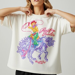 K&ouml;nnte beinhalten: Cremefarbenes T-Shirt mit einem farbenfrohen Aufdruck einer Cowgirl auf einem lila Pferd, mit dem Text "Rootin Tootin" und "Good Vibes Club". Die Cowgirl tr&auml;gt einen roten Hut, ein gelbes Hemd und gr&uuml;ne Hosen.