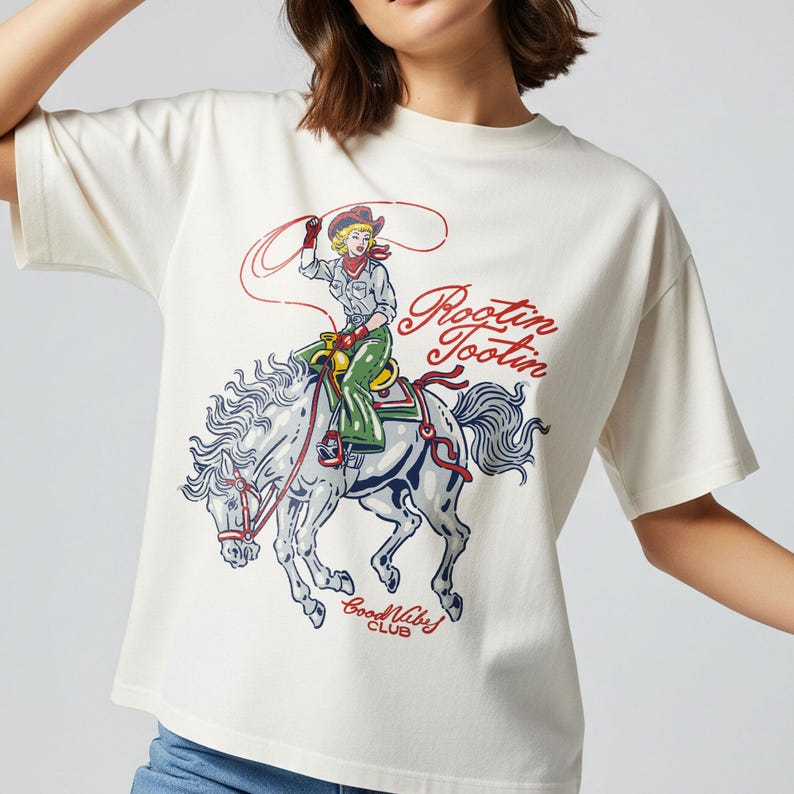 May include: Cream-colored t-shirt featuring a graphic of a cowgirl riding a horse with a lasso. The design includes the text "Rootin' Tootin'" in red script and "Good Vibes Club" in a smaller font. The cowgirl is wearing a cowboy hat and green pants.