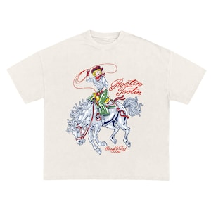 May include: Cream-colored t-shirt featuring a vintage-style illustration of a cowgirl riding a bucking horse. The cowgirl is wearing a red bandana and green pants, holding a lasso. The text "Rootin' Tootin" and "Good Vibes Club" are also on the shirt.