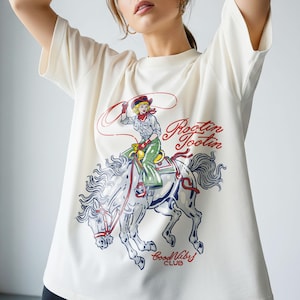 May include: Cream-colored t-shirt featuring a graphic of a cowgirl riding a horse with a lasso. The design includes the text "Rootin' Tootin" in red script and "Good Vibes Club" in blue. The cowgirl wears a red bandana and hat.