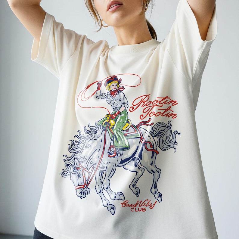 May include: Cream-colored t-shirt featuring a graphic of a cowgirl riding a horse with a lasso. The design includes the text "Rootin' Tootin" in red script and "Good Vibes Club" in blue. The cowgirl wears a red bandana and hat.