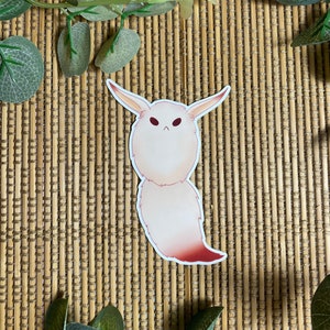 May include: A white and pink fuzzy creature with large ears and a red tail. The creature has a grumpy expression on its face. The creature is a sticker on a brown woven background.