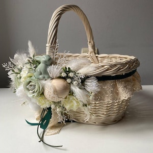 Puede incluir: Una cesta de mimbre blanca con un conejito verde y flores blancas, decorada con encaje y una cinta verde. La cesta es perfecta para Pascua.